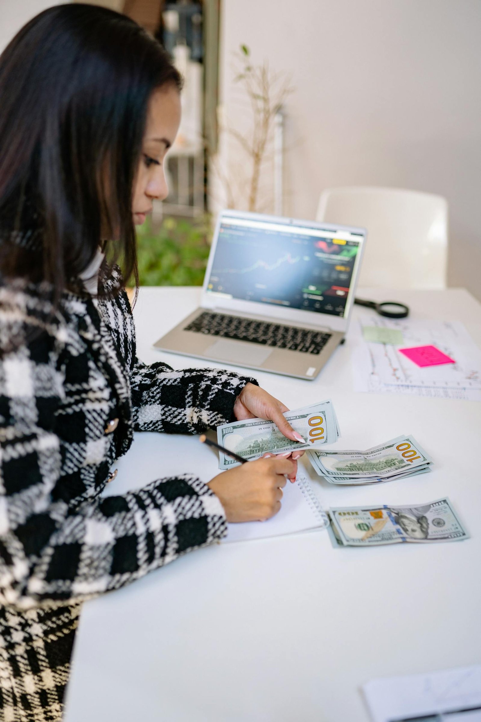 A woman focuses on counting dollar bills at a table with a laptop showing stock charts.