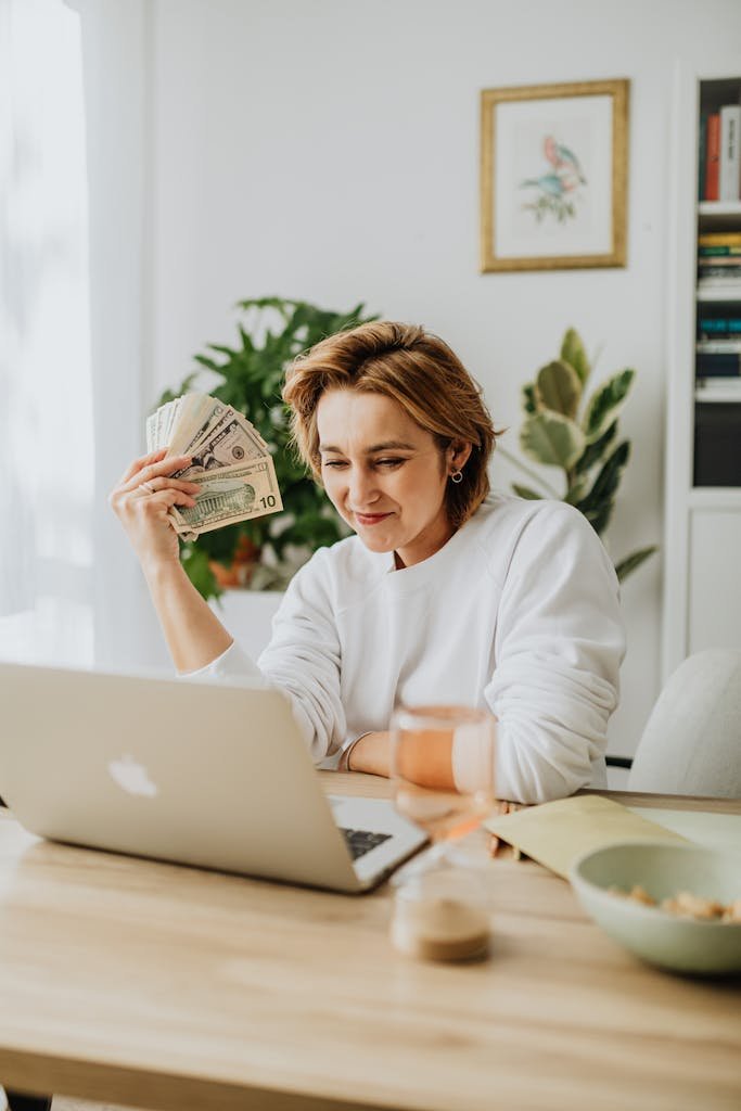 Woman seated at a desk holding dollar bills, working on a laptop, showcasing financial success from home.