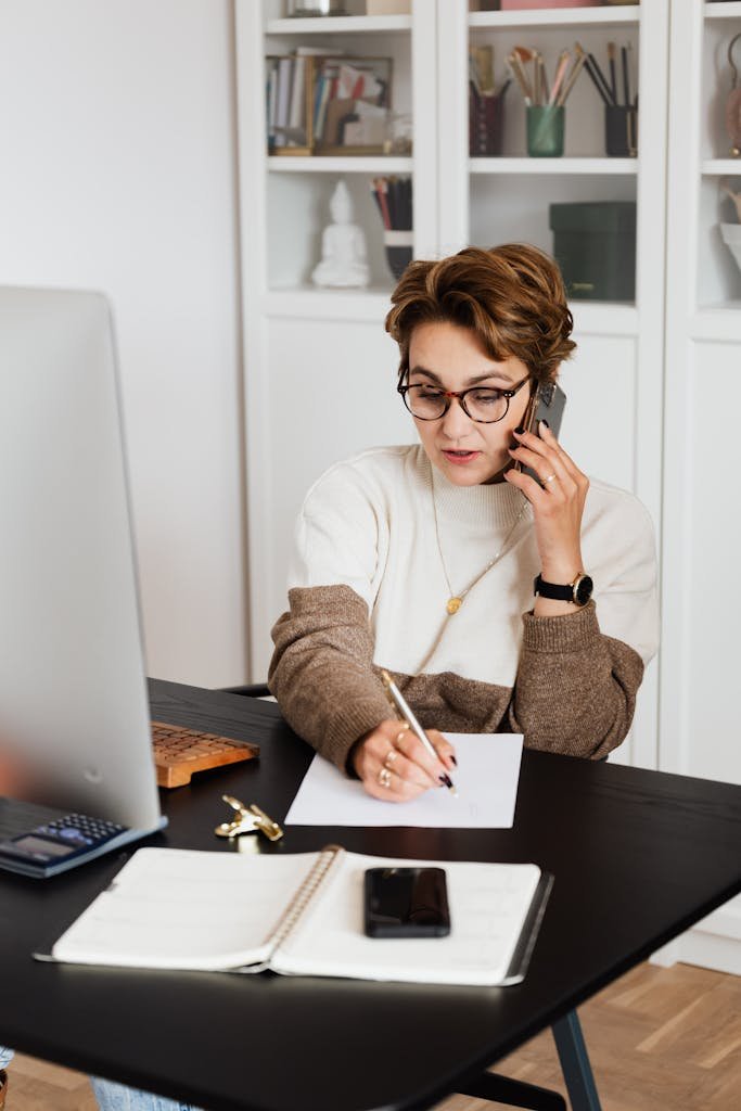 A woman working on her budget remotely from home, engaging in a phone call while taking notes.