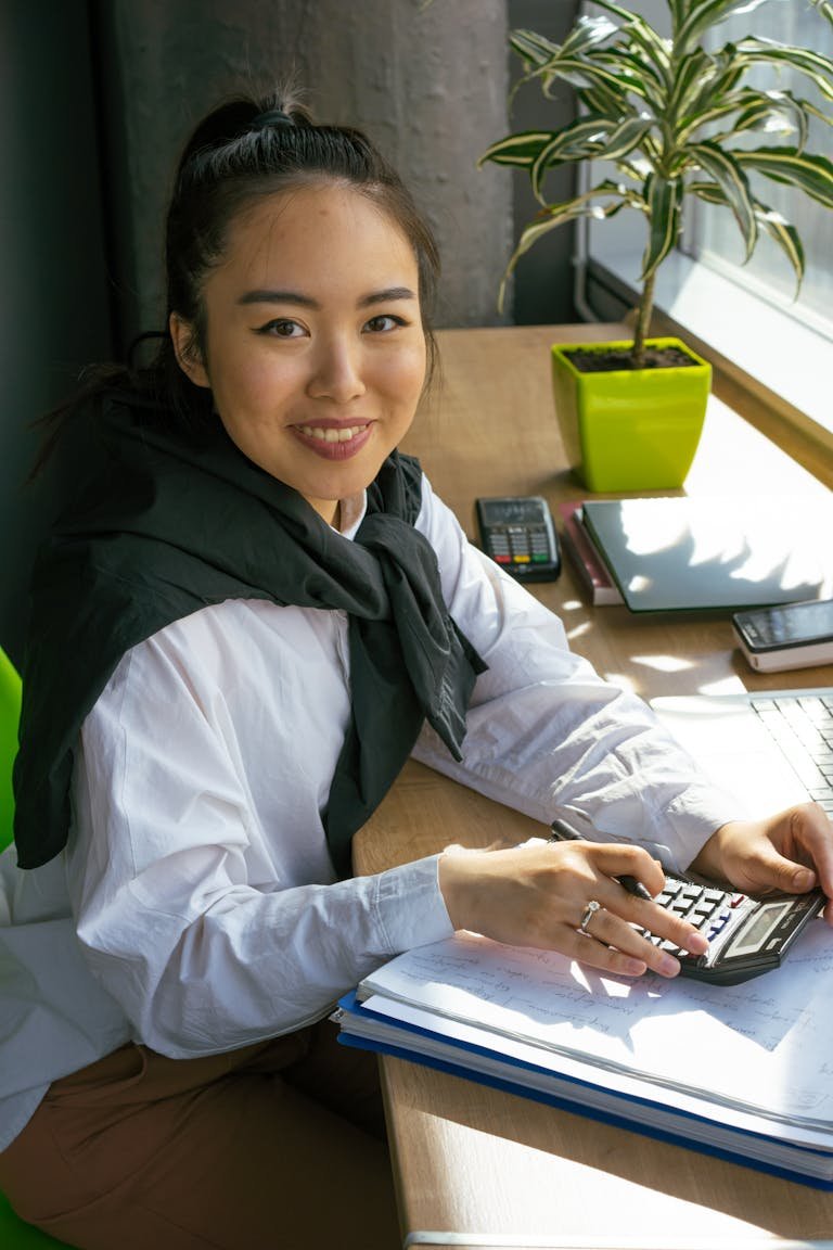 Asian woman working with calculator and laptop in a sunny office, smiling and focused.
