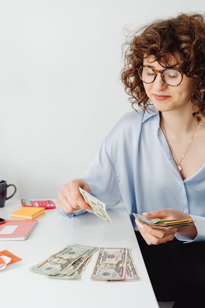 Caucasian woman in blue top counting money at desk with eyeglasses and a necklace. Personal finance strategies for women