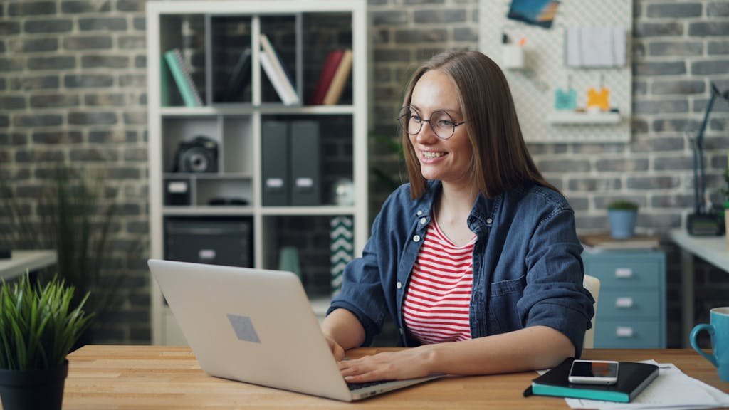 Cheerful woman working on laptop in modern office setting with smartphone and notebook.