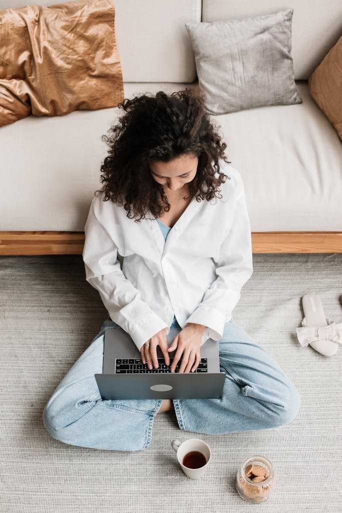 Top view of woman sitting on the floor, typing on a laptop with tea and cookies nearby.