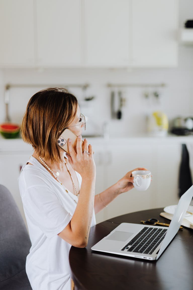 Woman multitasking with a smartphone and laptop while drinking coffee in a cozy home setting working on her budget.