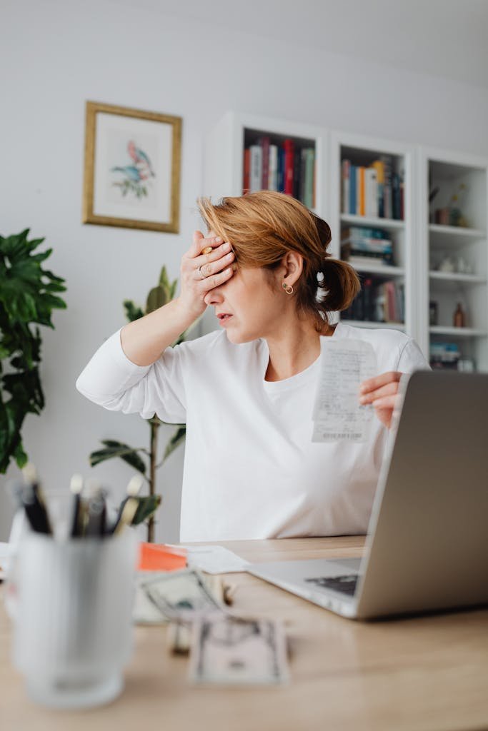 Worried woman counting expenses on a laptop while holding a receipt at her desk. Debt shame cycle