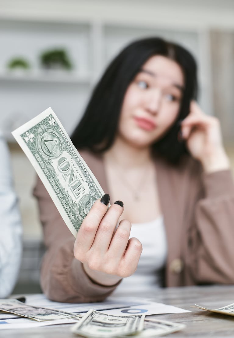 Young woman with dark hair holding a one-dollar bill, seated at a table.
