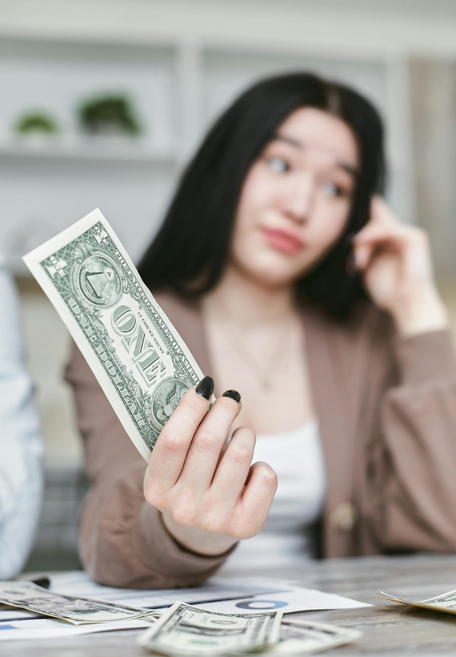 Young woman with dark hair holding a one-dollar bill, seated at a table.