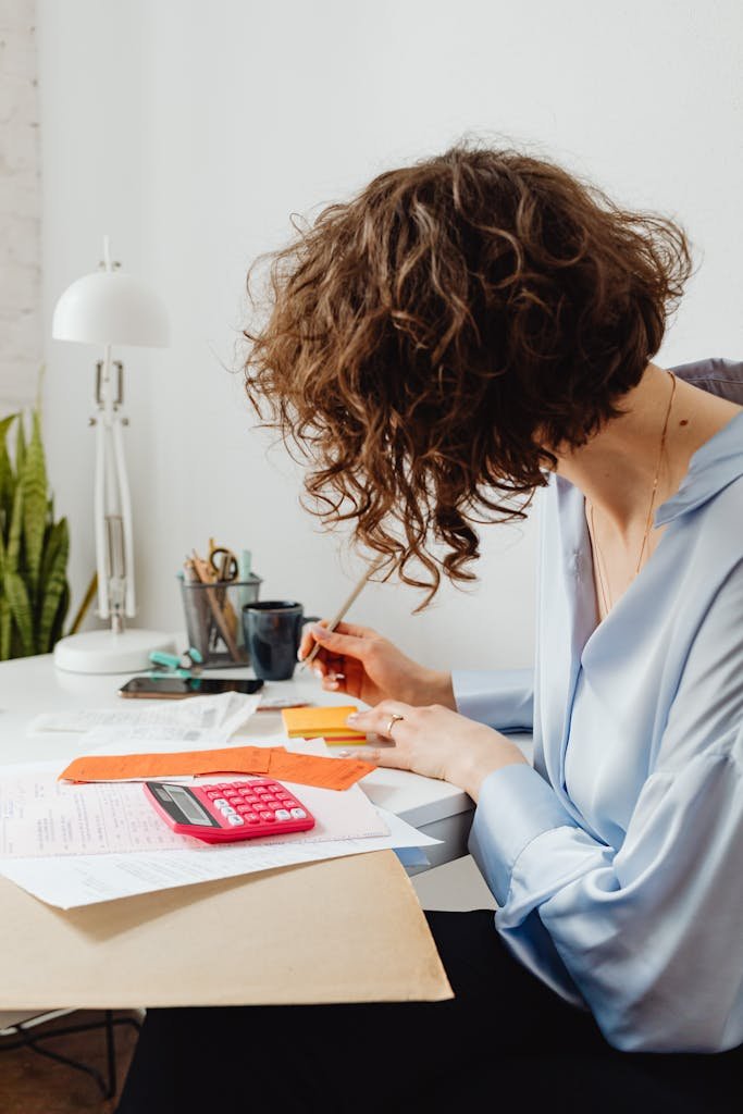 A woman in a blue blouse writing at a desk with a calculator and documents.