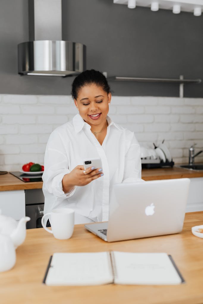 A woman using her smartphone while working on a laptop in a bright, modern kitchen setting.