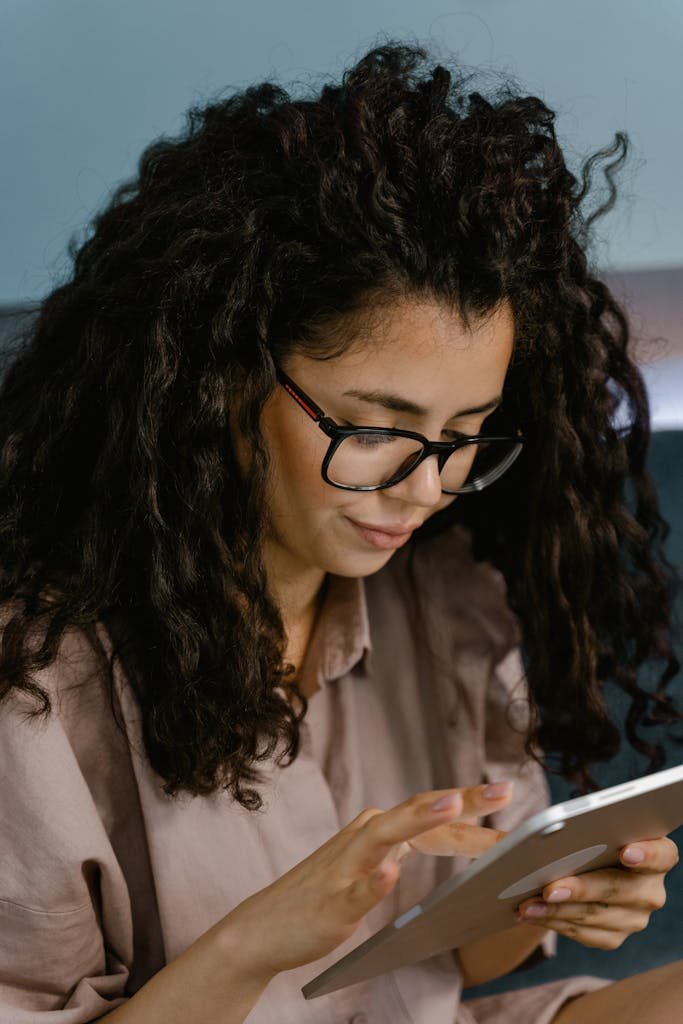 A woman with curly hair and eyeglasses using a tablet indoors, focusing intently on her work.