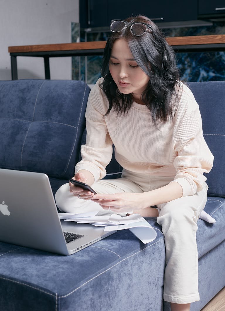 A young woman sits on a sofa managing her bills with a laptop and smartphone.
