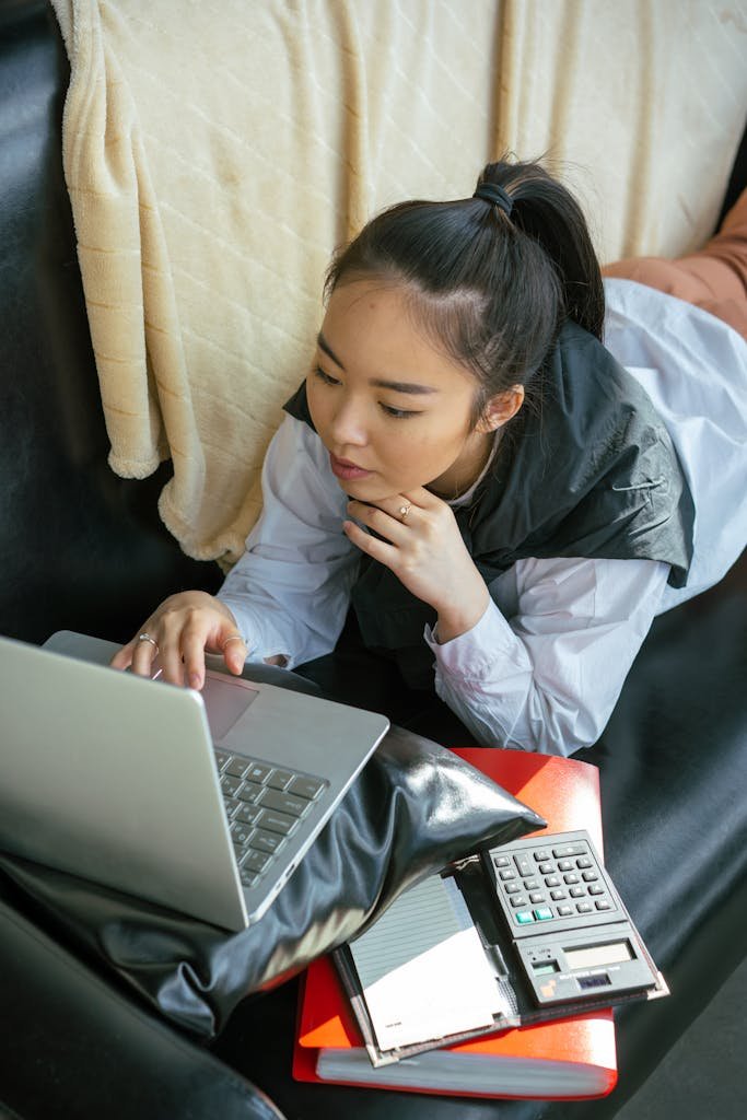 Asian woman lying on couch using laptop, focused on work. Cozy indoor setting.