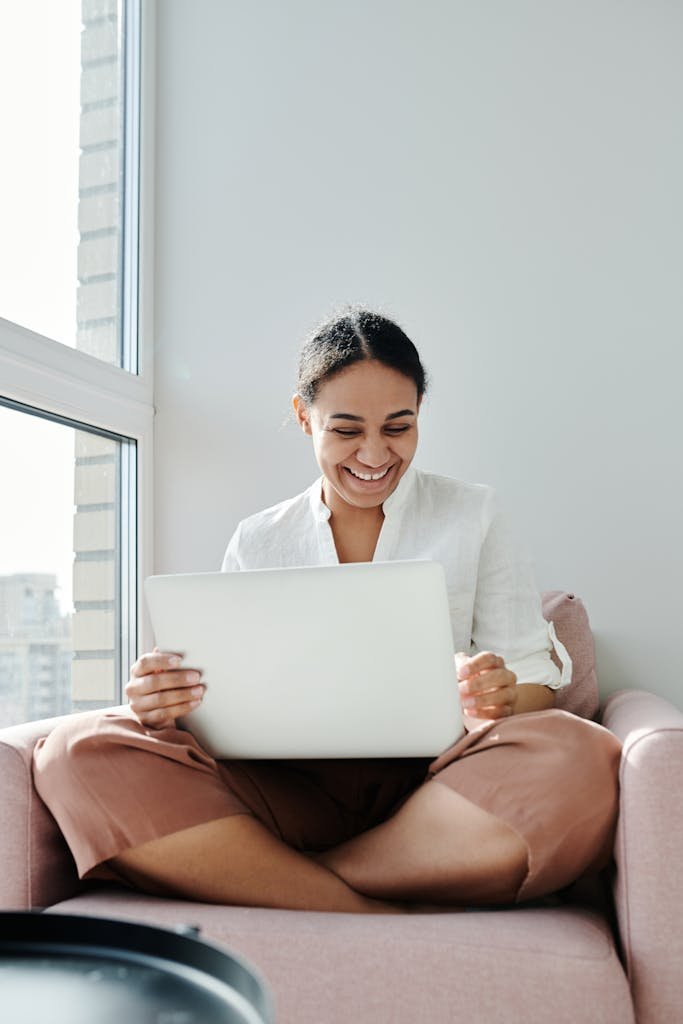 Cheerful woman using a laptop while sitting cross-legged on an armchair by a sunny window. High-yield savings account