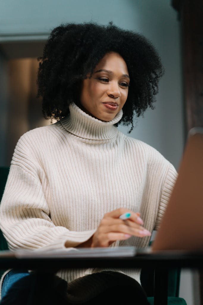 Happy woman in sweater working on laptop with curly hair, indoors during the day.