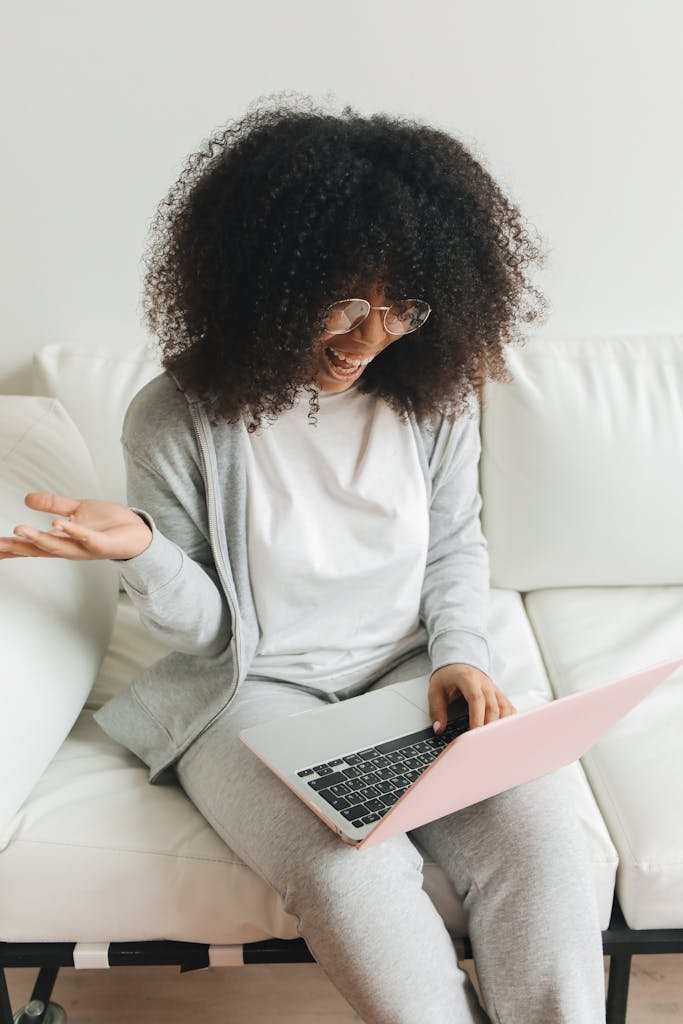 Smiling woman with curly hair working on a laptop while sitting on a white sofa indoors.