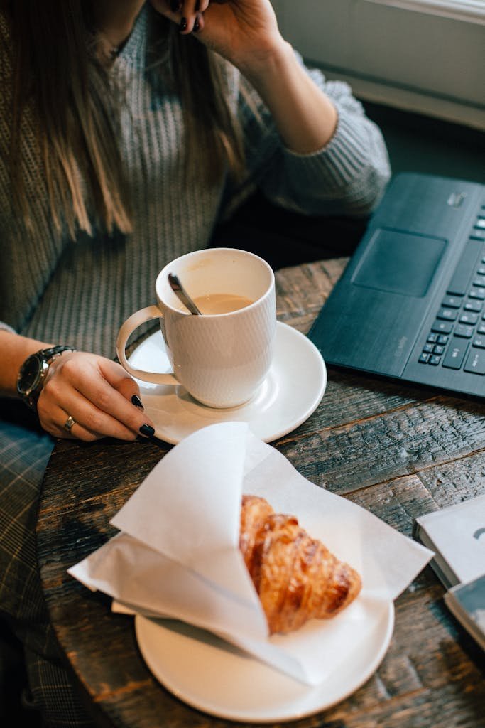 Warm café atmosphere with coffee, croissant, and casual laptop work setting.