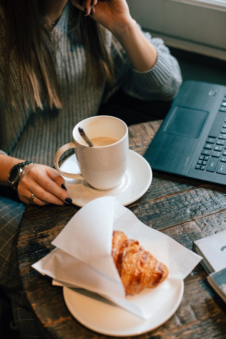 Warm café atmosphere with coffee, croissant, and casual laptop work setting.