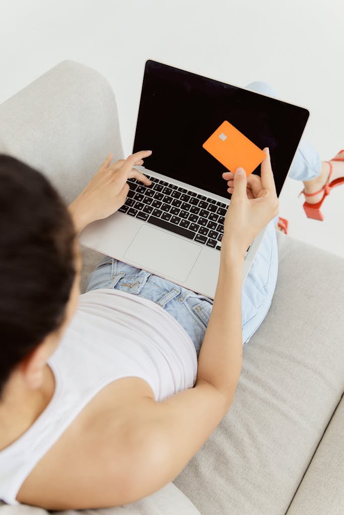 Woman holding orange credit card while shopping online on laptop from a sofa. Lifestyle creep