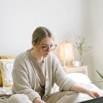 Woman in casual attire working on a laptop from a cozy bedroom setting, emphasizing remote work lifestyle. 20-Minute Money Habit