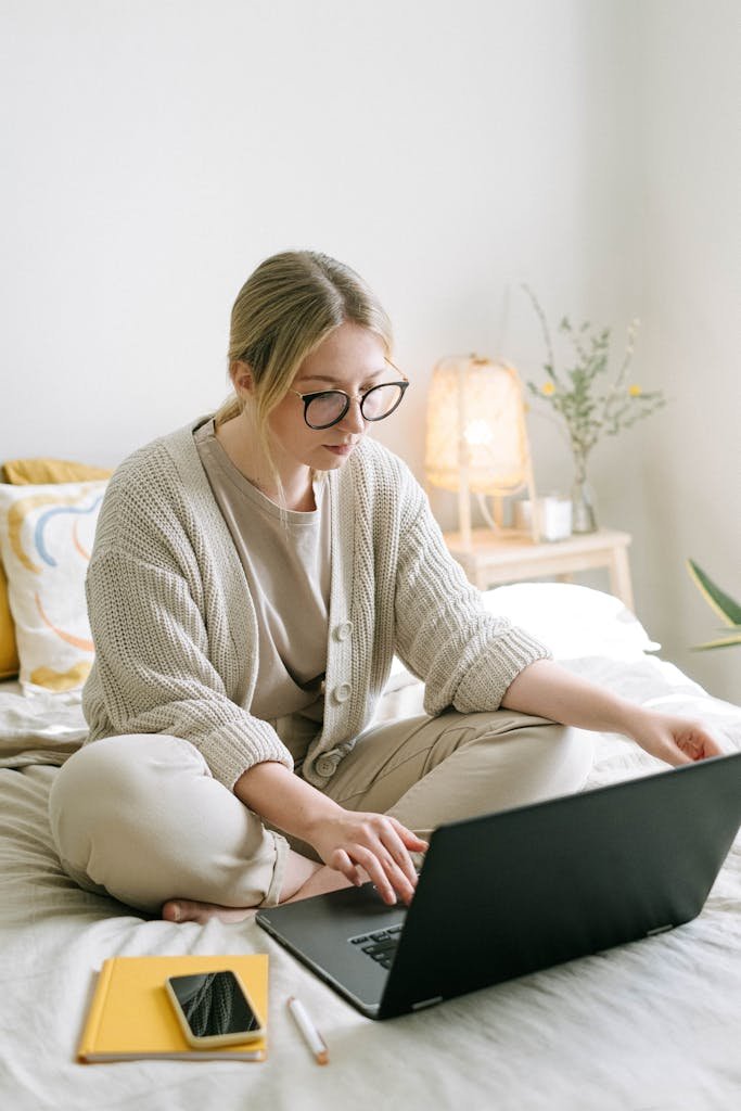 Woman in casual attire working on a laptop from a cozy bedroom setting, emphasizing remote work lifestyle.