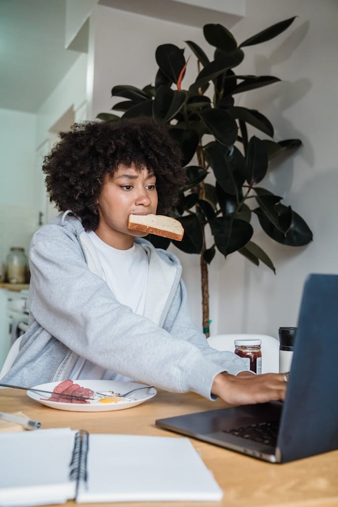 Woman multitasks with breakfast and laptop work in a cozy home setting.