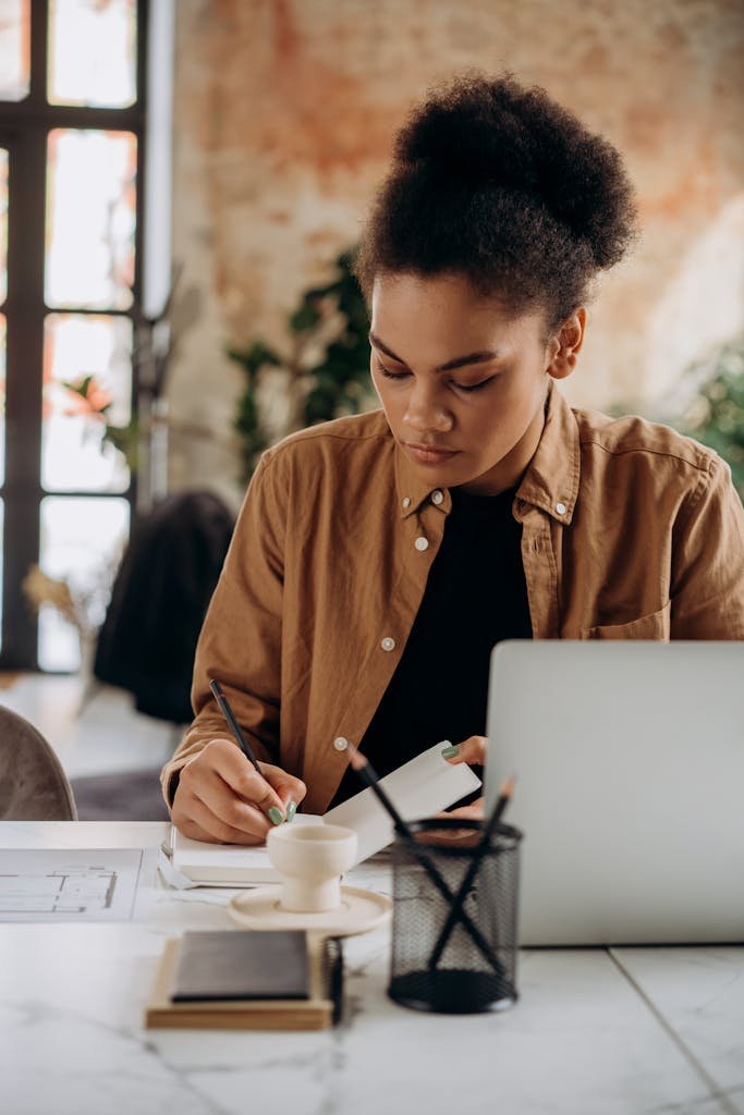 Woman taking notes at a desk with a laptop, focusing on her tasks in a cozy workspace.
