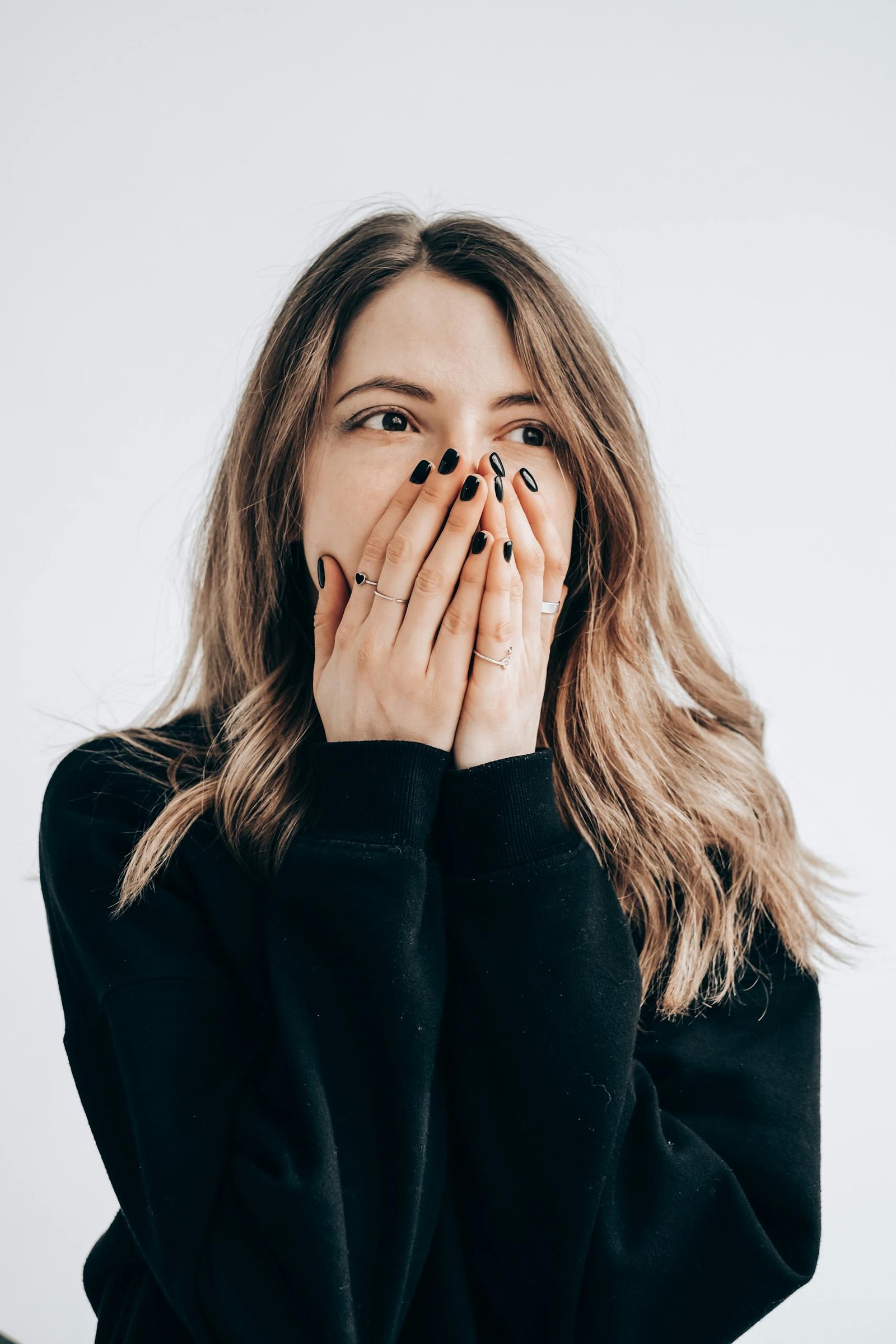 A surprised young woman covers her mouth with her hands, wearing a black sweater.