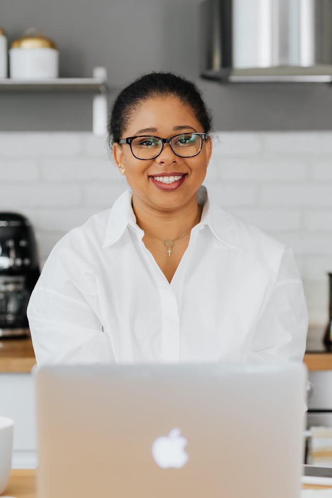 A happy woman in a white shirt and glasses working on a laptop at home.