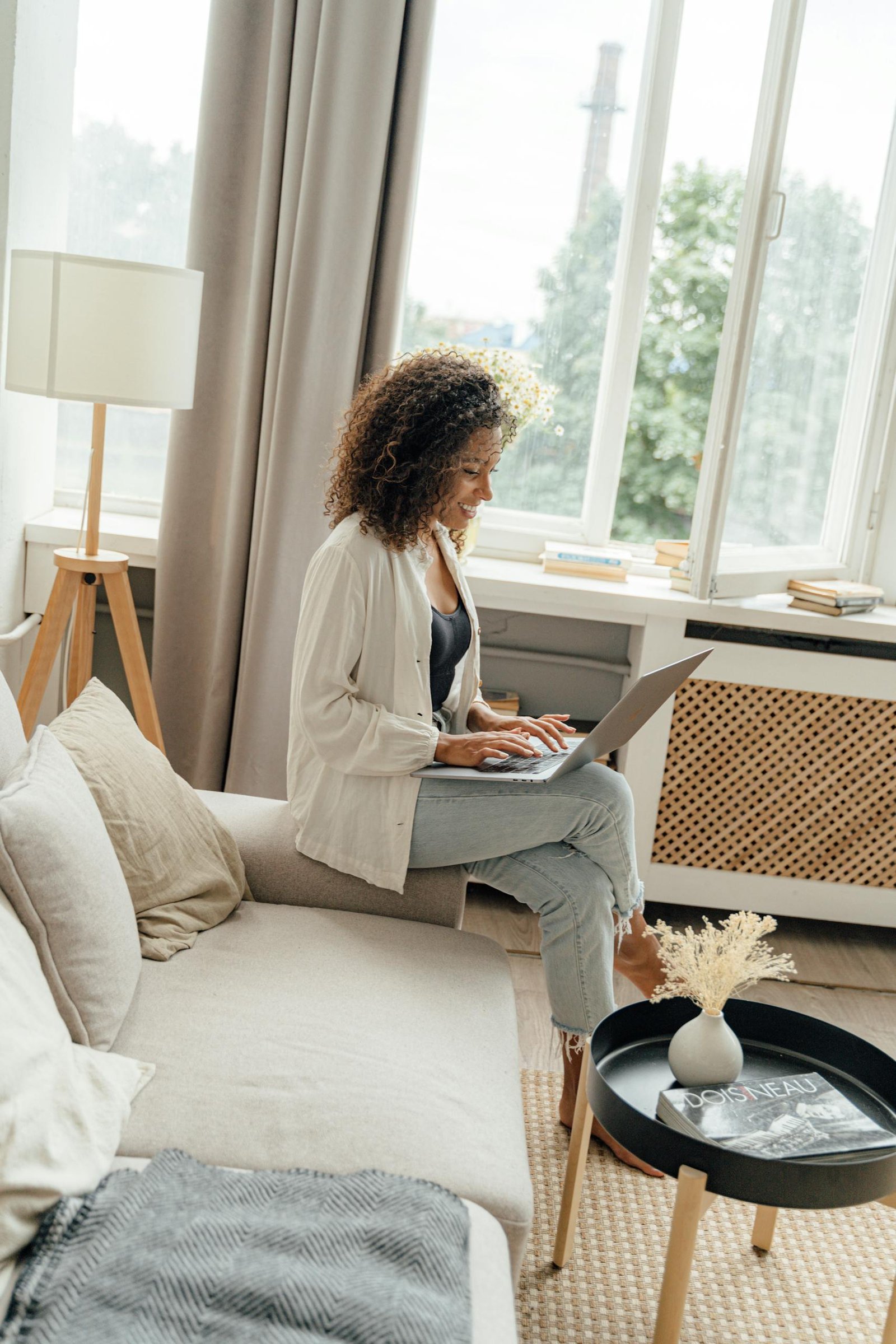 A woman sitting comfortably on a sofa using a laptop in a bright, cozy living room.