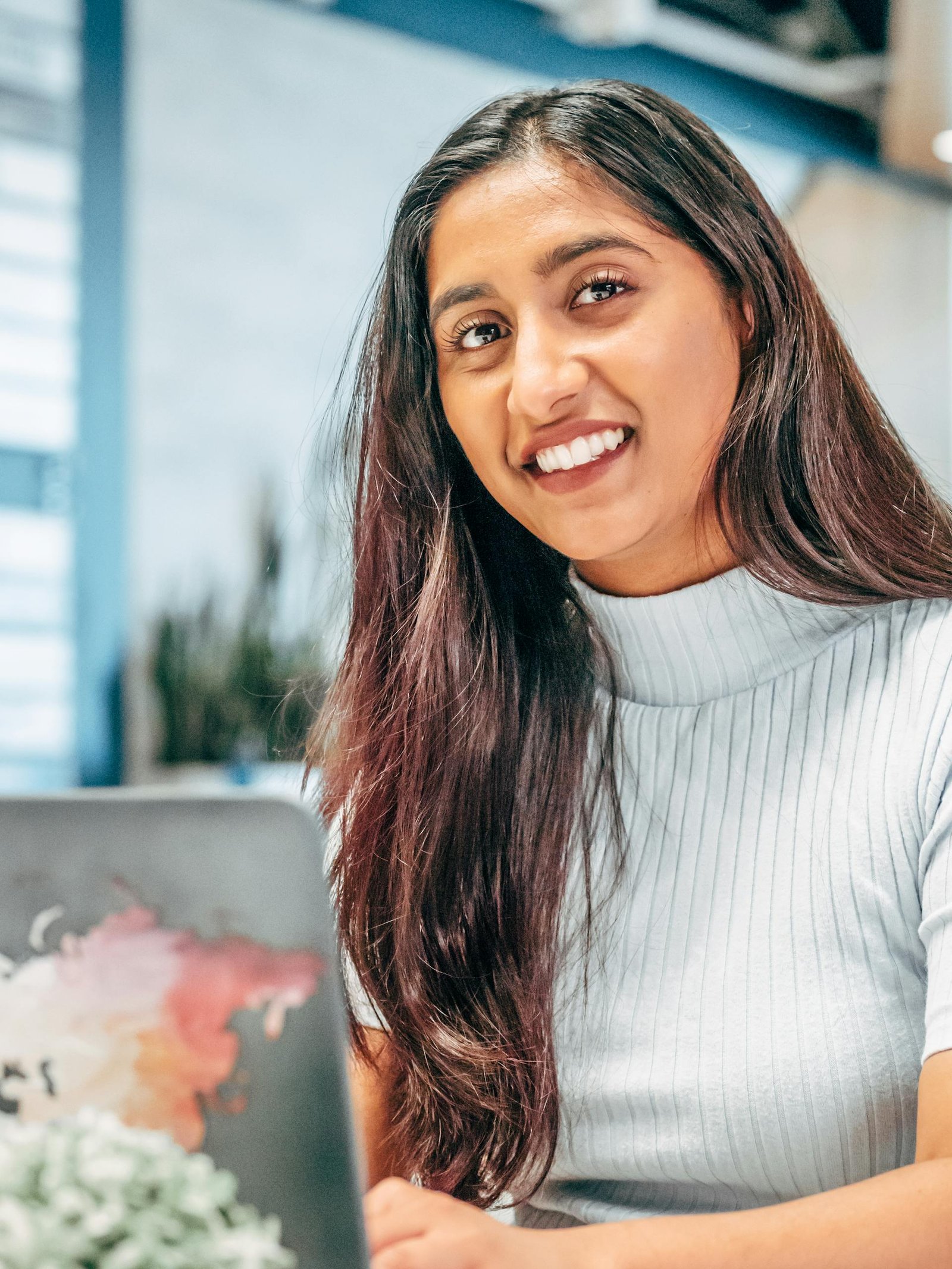 Portrait of a woman with long hair smiling at a laptop indoors, conveying positivity.