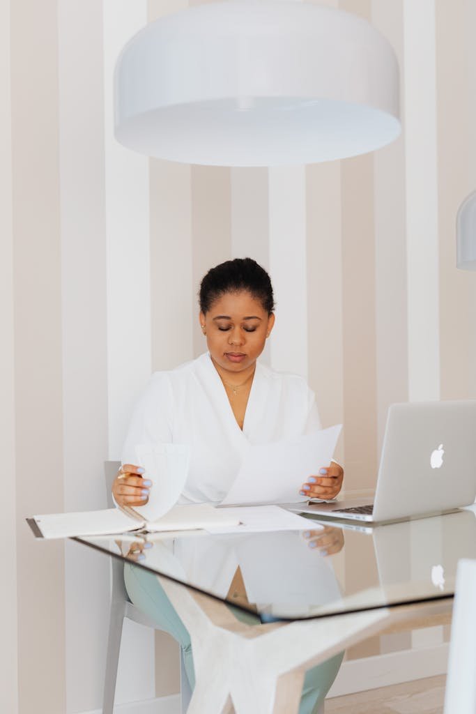A black woman in a white top focused on work with documents and laptop in a stylish office.