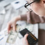 A woman calculates finances using a calculator with banknotes and documents on the table.