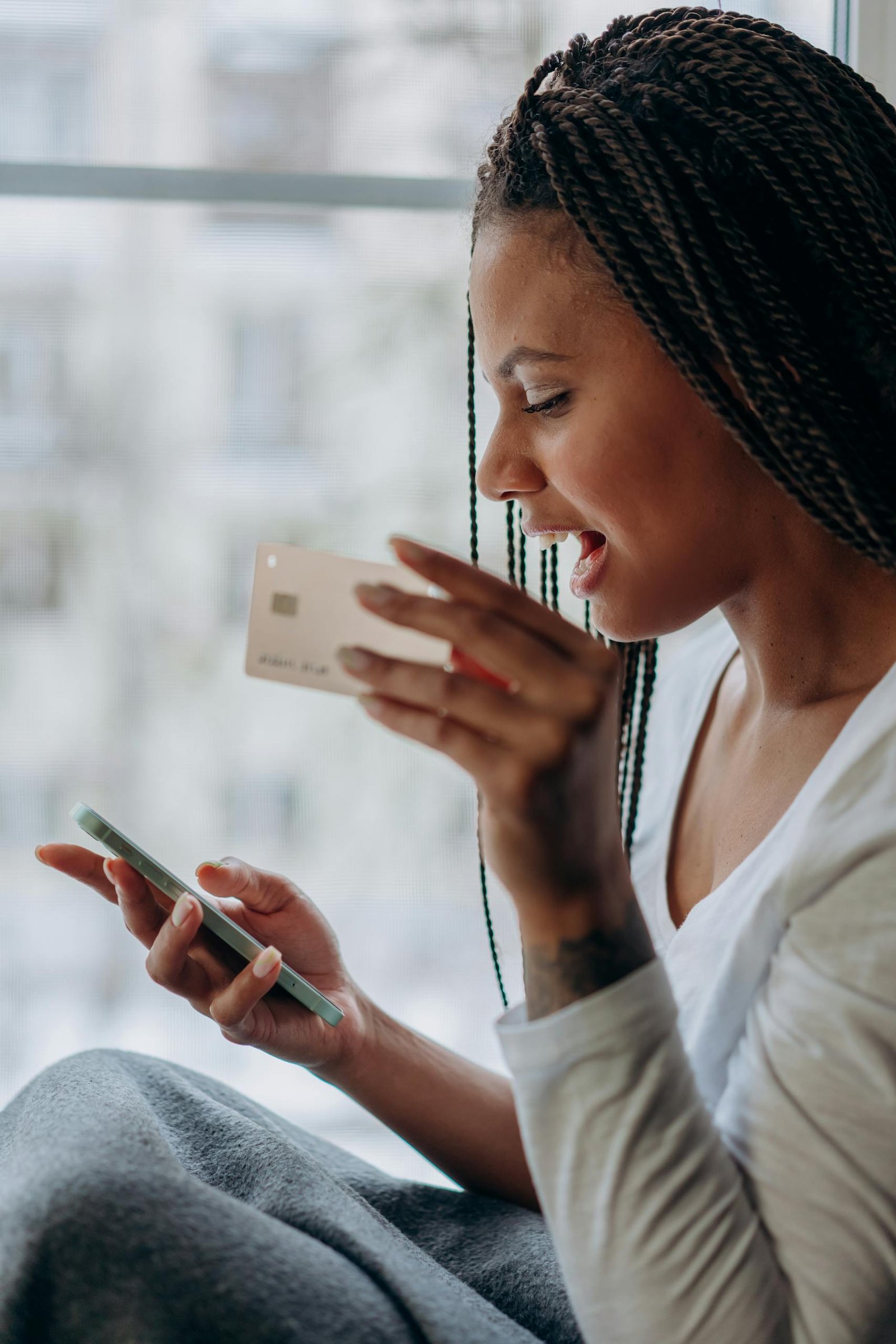A woman smiles while shopping online using her smartphone and credit card by a window.