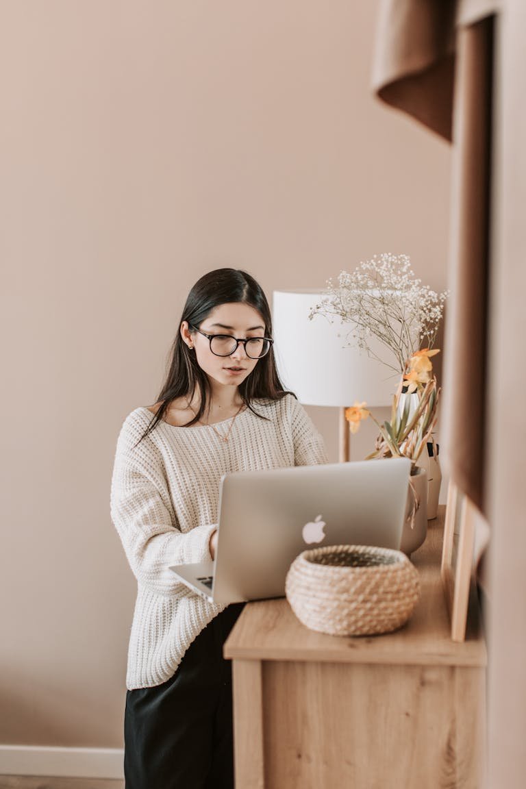Concentrated young female in trendy casual outfit using laptop while standing near cabinet in cozy living room of modern apartment
