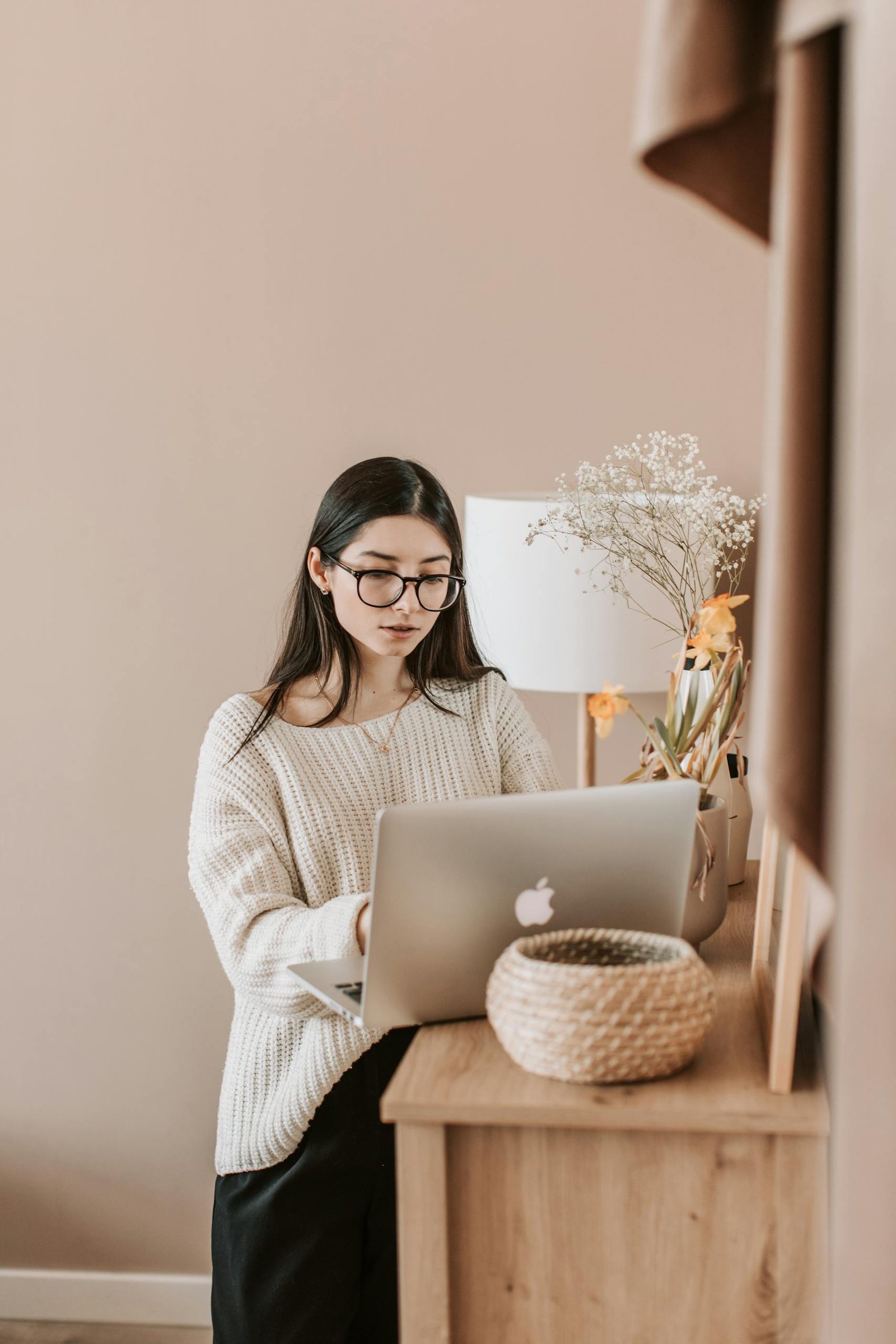 Concentrated young female in trendy casual outfit using laptop while standing near cabinet in cozy living room of modern apartment