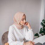 Woman wearing a hijab working on a laptop at a desk in a minimalist room with a bottle and notebook.