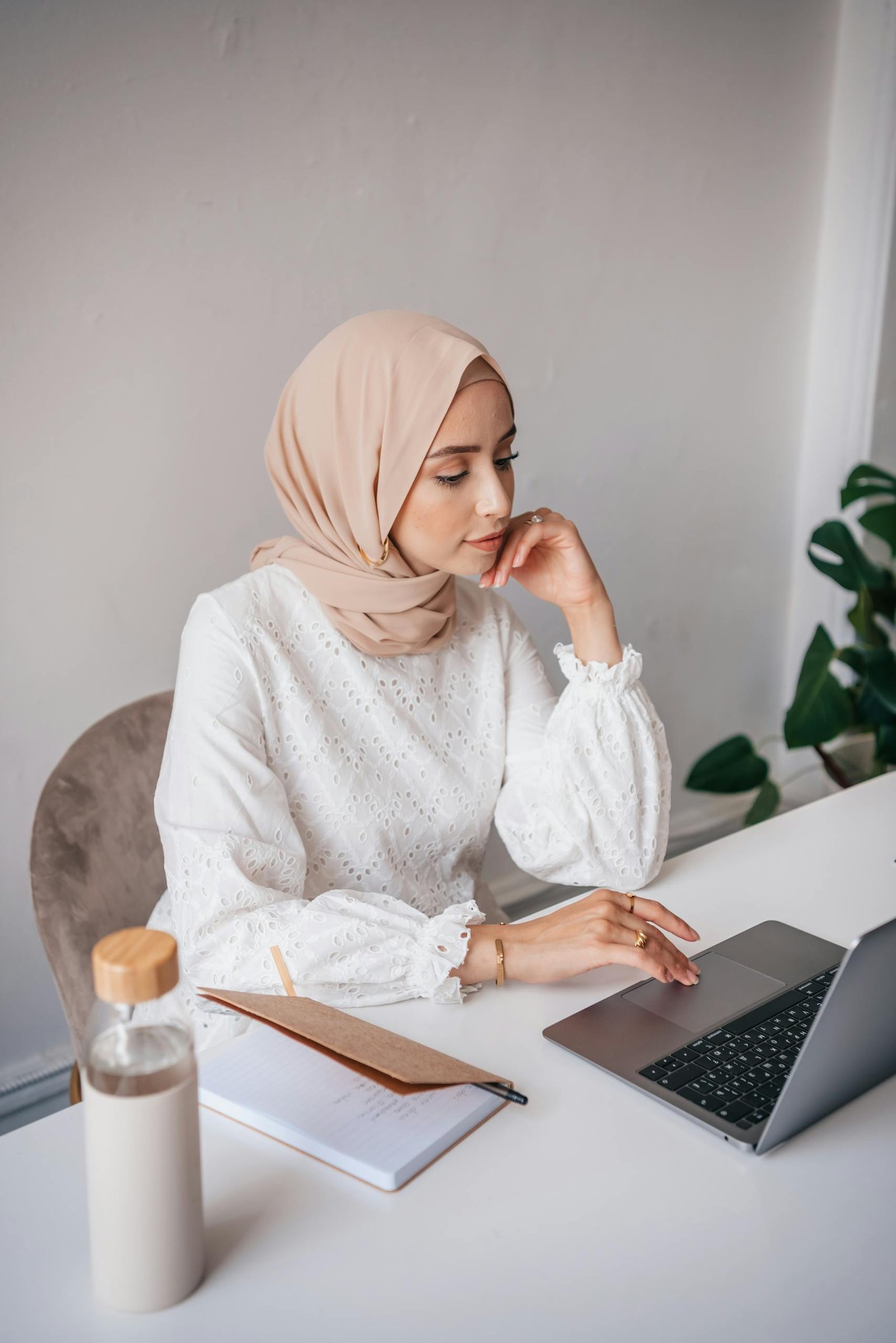 Woman wearing a hijab working on a laptop at a desk in a minimalist room with a bottle and notebook.
