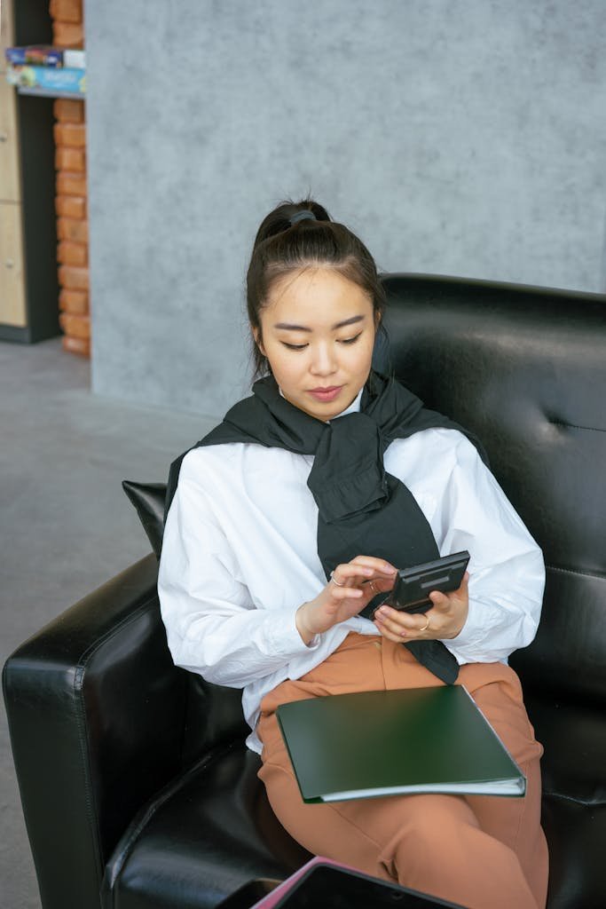 Asian woman sitting on sofa using calculator while relaxing indoors.