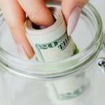 Hand placing rolled US dollars into a glass jar, representing savings and financial planning.