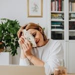 Happy woman sitting at a desk with cash and using a laptop, in a cozy home office setting.