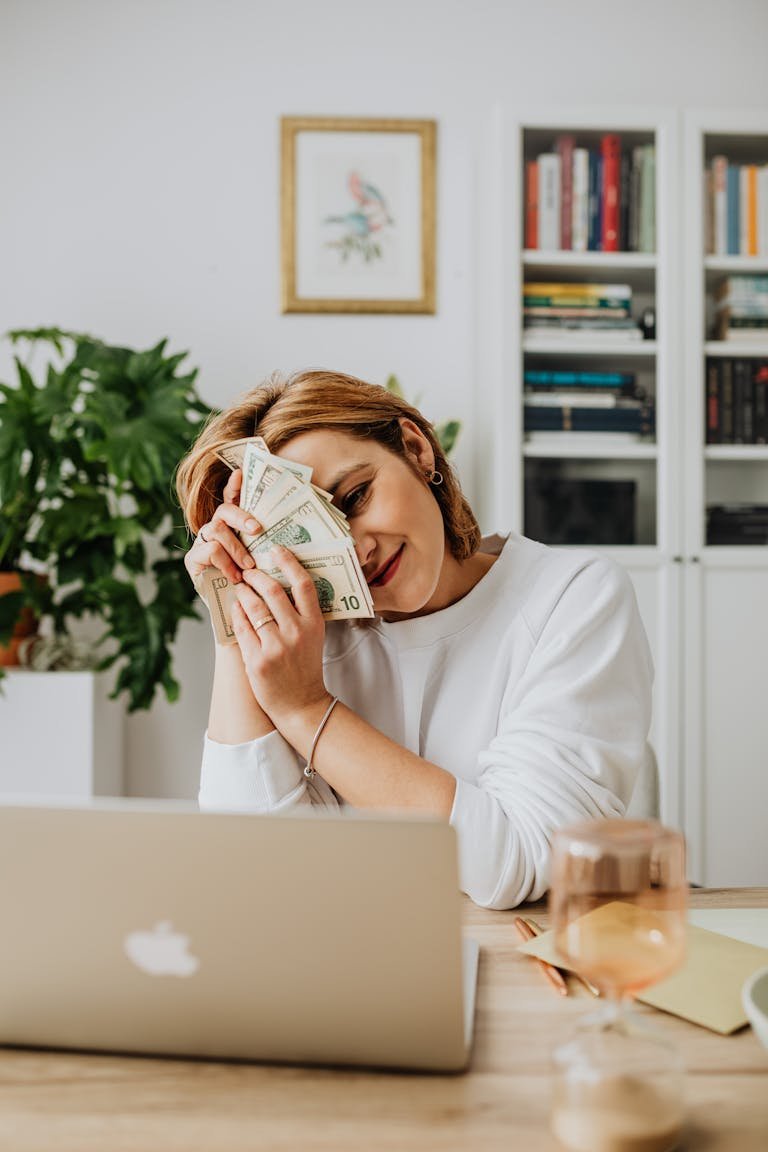 Happy woman sitting at a desk with cash and using a laptop, in a cozy home office setting.