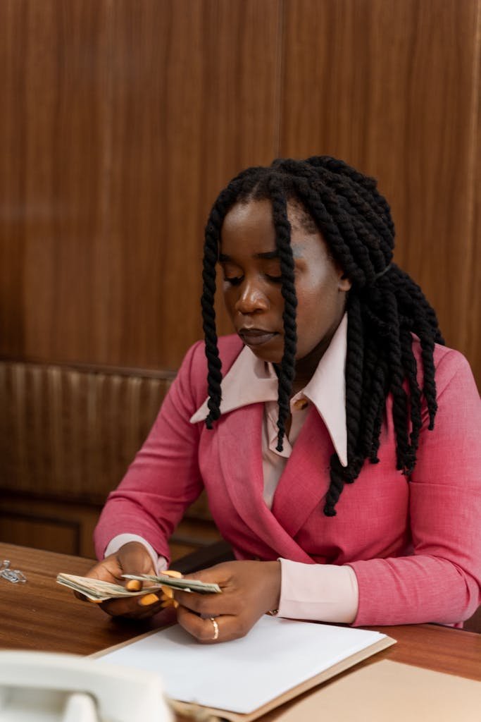 African American woman in pink blazer counts money at the desk with focused expression.