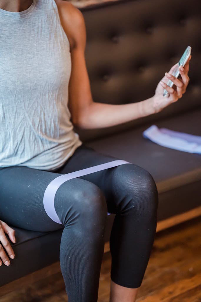 Faceless sportswoman in activewear sitting on couch and surfing internet on smartphone while exercising with elastic resistance rope on legs during training