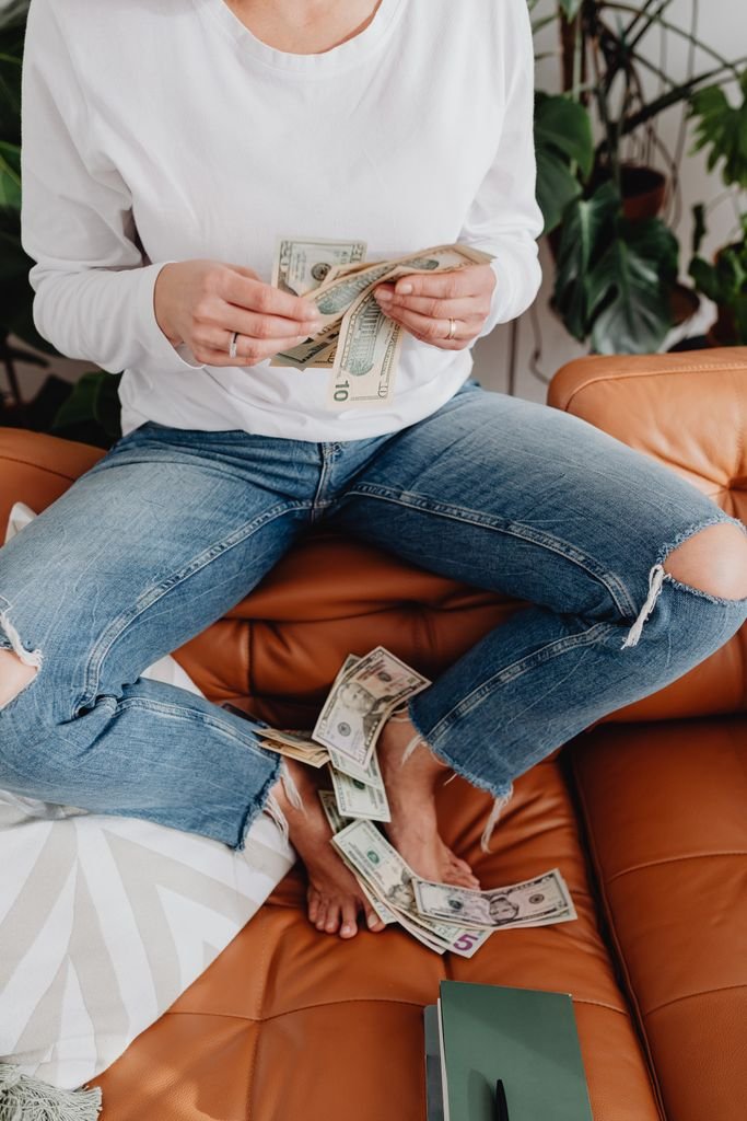Casual scene of a woman counting dollar bills on a sofa surrounded by plants.