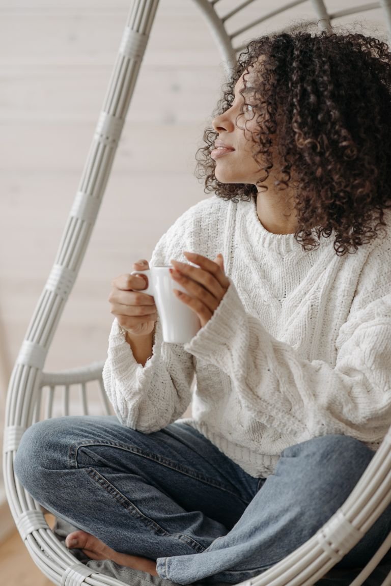 An African American woman with curly hair enjoys coffee in a cozy indoor setting.