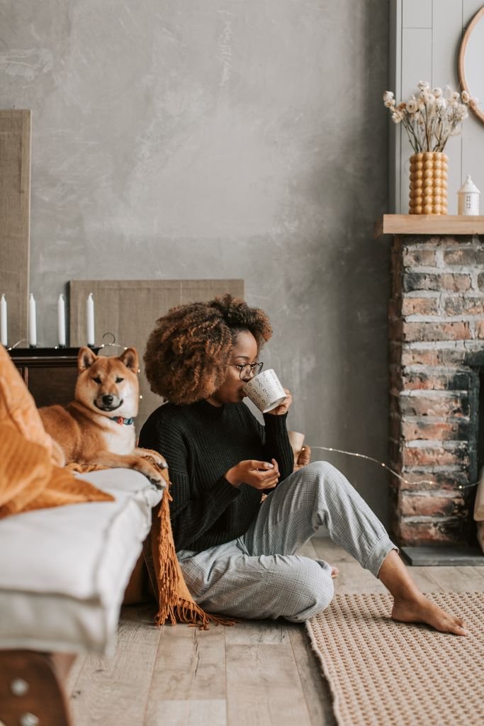 Relaxing at home, an African American woman enjoys coffee beside her pet dog in a cozy living room.